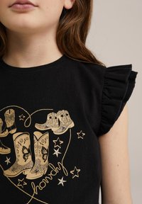 Juodas medvilnės marškinėliai trumpomis plunksnomis rankovėmis; puošia auksinė grafika su kaubojaus batais ir žvaigždėmis, akcentuojanti dekoratyvinius elementus.