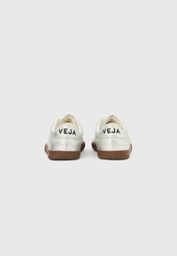 Coppia di sneakers bianche Veja con suole marroni viste da dietro, che mostrano il logo "VEJA" su ogni linguetta del tallone.