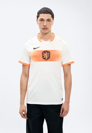 Νέος άνδρας με κοντά μαύρα μαλλιά, ντυμένος με λευκό και πορτοκαλί αθλητικό μπλουζάκι με έμβλημα λιονταριού και μαύρο παντελόνι, στέκεται μπροστά σε ένα απλό λευκό φόντο.