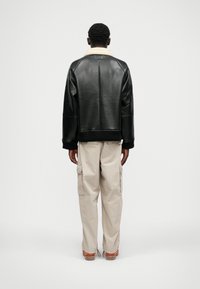 Czarna skórzana kurtka z kremowym kołnierzem z owczej wełny, luźny fason, ściągacze na mankietach i u dołu, w połączeniu z beżowymi spodniami cargo i brązowymi butami.