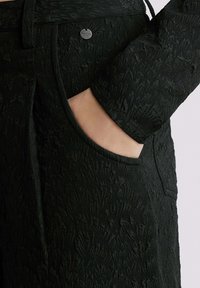 Zwarte textuur broek met een subtiel patroon, voorzien van een zijzak en een metalen merkembleem. De stof lijkt geweven en heeft een matte afwerking.