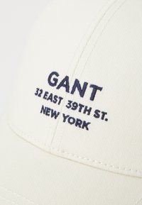 Vit basebollkeps med marinblå broderad text: "GANT," "32 EAST 39TH ST.," och "NEW YORK." Har en böjd skärm och texturerad tyg.