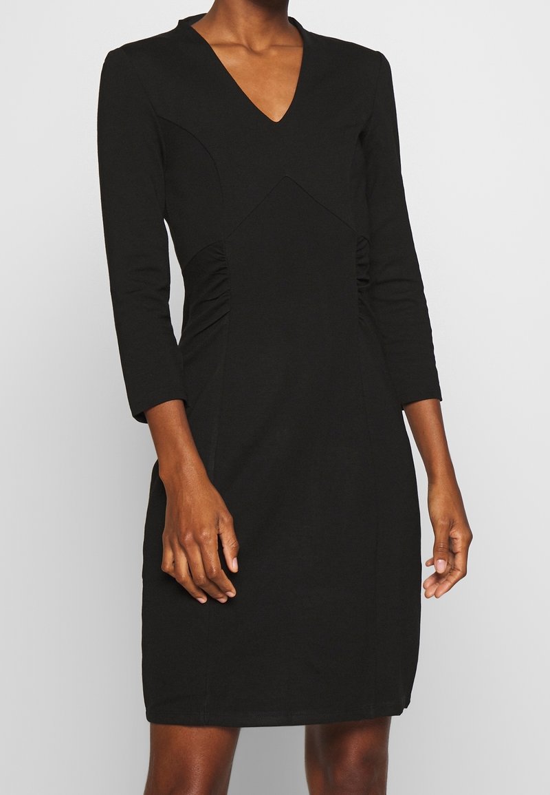 Robe noire à manches longues avec un décolleté en V, dotée de fronces sur les côtés et d'une silhouette ajustée, fabriquée à partir d'un tissu lisse et extensible.
