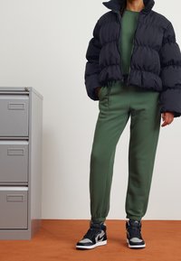 Svart puffjacka med ribbad krage, grön sweatshirt och matchande tapered joggers. Svart och grå högsko sneakers fulländar outfiten.
