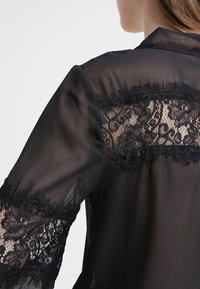 Czarna przezroczysta bluzka z koronkowymi zdobieniami na rękawach i plecach, z delikatnym kwiatowym wzorem i miękką, zwiewną fakturą.