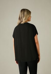 Svart kortärmad topp med en texturerad design, lös passform och rundad nederkant, i kombination med svarta byxor. Utsikt bakifrån.