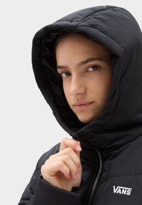 Chaqueta de plumas negra con capucha, que incluye un cierre de cremallera y puños elásticos. El material parece suave e insulated, con un logotipo visible.