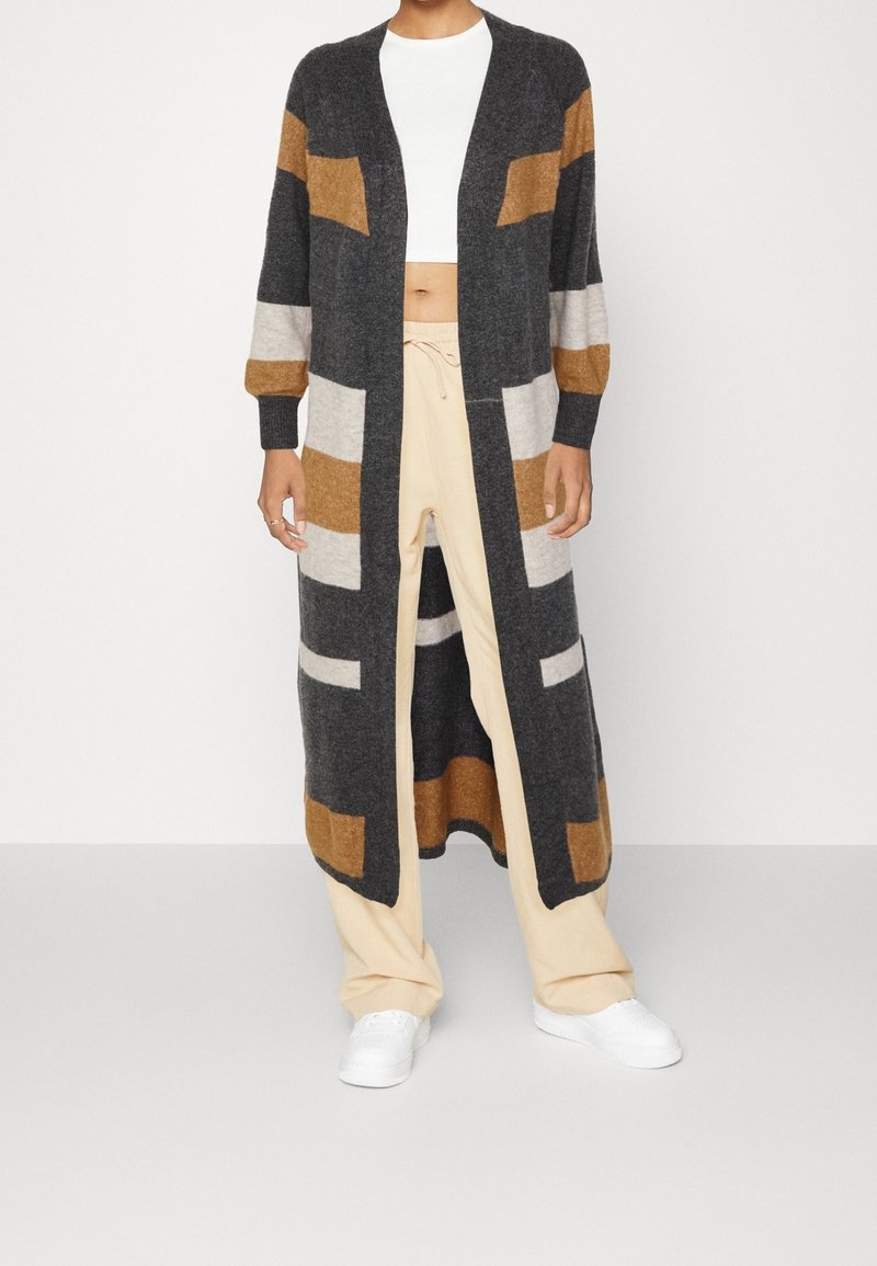 Personne portant un cardigan long ouvert devant avec des blocs géométriques gris, marron et beige, un pantalon beige, un crop top blanc et des baskets blanches.