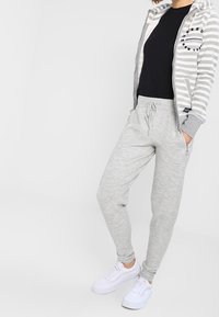 Persona che indossa una felpa grigia a righe con zip, una maglietta nera, pantaloni jogger grigio chiaro e sneakers bianche, in piedi con le mani in tasca.