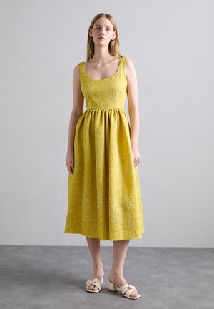 Yellow sleeveless dress with floral embroidery, fitted bodice, and full skirt. Paired with white block-heeled sandals.
