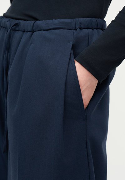 Χέρι στην τσέπη μπλε σκούρου, ελαστικής μέσης καθημερινών παντελονιών που φοριούνται με μαύρο μακρυμάνικο τοπ, εμφανιζόμενα από τη μέση μέχρι το μέσο του μηρού.