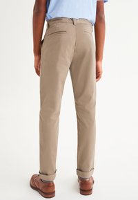 Beige Baumwollhosen mit zwei Gesäßtaschen, schmaler Passform und umgeschlagenen Bündchen, kombiniert mit braunen Lederschuhen und gestreiften Socken.