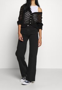 Mujer vestida con pantalones vaqueros de pierna ancha negros, un cárdigan de punto abierto negro sobre una camiseta blanca, y zapatillas negra y blancas, de pie contra un fondo liso.