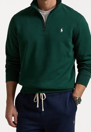 Mand iført mørkegrøn kvart-zip sweatshirt og marineblå bukser med snøre, med ur på venstre håndled.