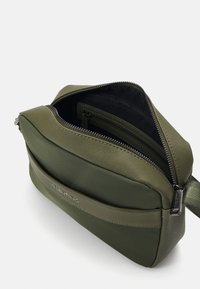 Grønn nylon crossbody-veske med glidelåslukking, ekstern lomme og sølvfarget logo. Teksturert overflate og justerbar stropp.