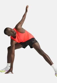En atletisk mand strækker sig i en lys koralfarvet tank top og sorte shorts med skrå snit. Han har hvide sokker og multifarvede sneakers på.