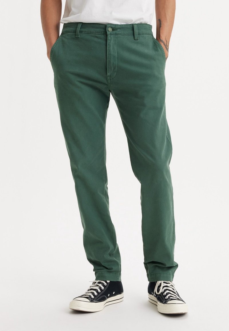 Chinos en coton verts avec une coupe droite, dotés d'une fermeture à bouton, de poches latérales, associés à des baskets montantes noires.