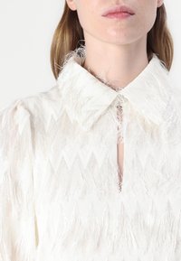 Robe blanche en tissu texturé avec un motif en zigzag et des détails à franges. Le col est pointu, complétant ainsi le design général.