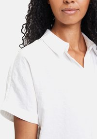 Weißes Leinenhemd mit einer entspannten Silhouette, kurzen Ärmeln und einem spitzen Kragen. Glatte Textur mit minimalen Details.