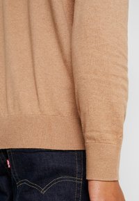 Maglione beige con orlo e polsini a coste, realizzato in un tessuto morbido, con una vestibilità leggermente ampia, abbinato a jeans in denim scuro.