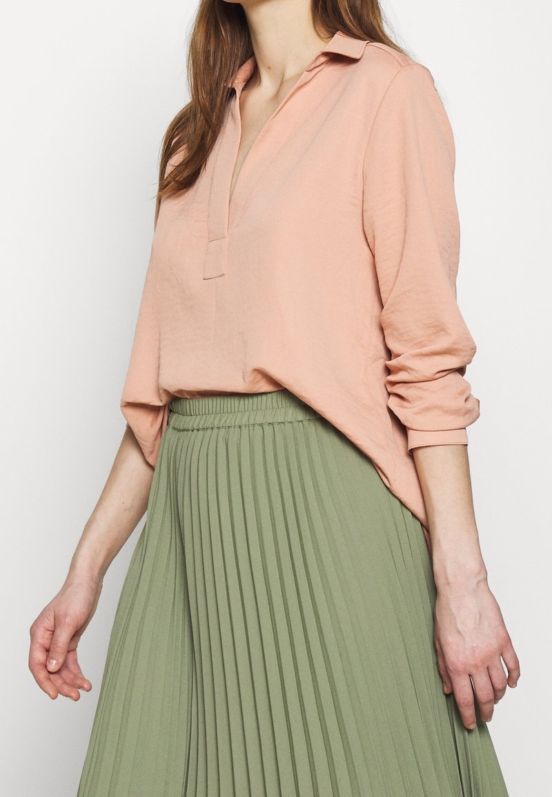 Femme portant un chemisier ample à manches longues couleur pêche, rentré dans une jupe plissée taille haute de couleur vert clair, sur un fond uni.