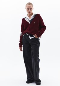Suéter de punto burdeos con detalle de trenzado y cuello blanco, superpuesto a una camisa blanca. Jeans negros de pierna ancha y zapatos negros.