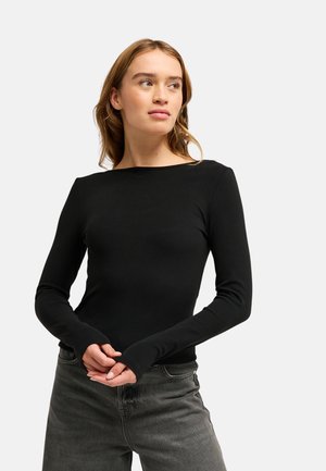 Jeune femme aux cheveux châtain clair portant un haut noir moulant à manches longues et un jean gris foncé, regardant vers sa droite.