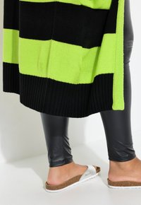 Cardigan lavorato a maglia con righe orizzontali nere e verdi neon, orlo a coste, abbinato a leggings neri lucidi e sandali bianchi con dettagli in sughero.