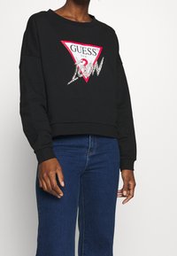 Felpa nera con design cropped, caratterizzata da un grande logo triangolare rosso con la scritta "GUESS" e dettagli scintillanti, abbinata a jeans blu.