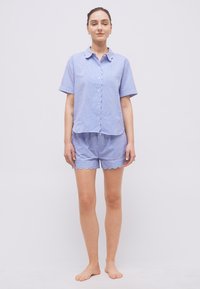 Γυναίκα όρθια, ξυπόλητη, φορώντας μπλε και λευκό ριγέ πουκάμισο με κοντά μανίκια και κουμπιά, και ταιριαστά σορτς με κυματιστά τελειώματα.