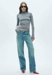 Šedý priliehavý rolák, svetlomodré široké nohavice a červené topánky na špičku. Model stojí pred jednoduchým bielym pozadím.