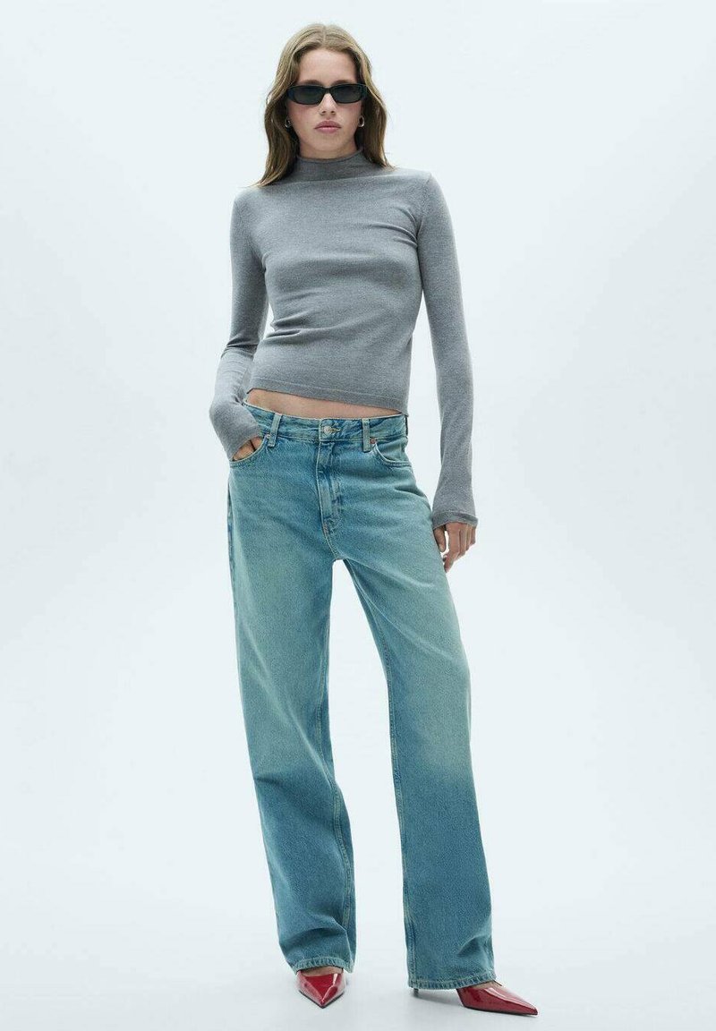 Šedý priliehavý rolák, svetlomodré široké nohavice a červené topánky na špičku. Model stojí pred jednoduchým bielym pozadím.