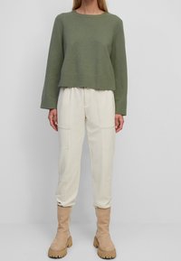 Pull court en maille vert olive à manches longues, associé à un pantalon crème fuselé avec poches avant, et des bottes beige montantes jusqu'au genou.