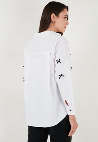 White long-sleeve shirt with black cross-stitch details on sleeves, button cuffs, and a curved hem. Made of a smooth, lightweight fabric.