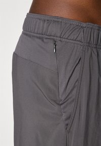 Harmaat urheilushortsit, joissa on joustava vyötärönauha, kuvioitu kangas, vetoketjullinen sivutasku ja kontrastiväriset sauman yksityiskohdat.