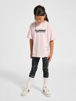 Junges Mädchen mit dunklen Haaren im Pferdeschwanz, trägt rosa übergroßes T-Shirt, schwarze gemusterte Leggings, weiße Socken und weiße Turnschuhe, steht und schaut nach unten.
