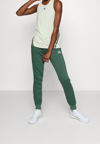 Débardeur de sport sans manches vert clair avec des rayures noires sur les côtés, associé à un pantalon de survêtement vert foncé fuselé et des baskets blanches avec des accents dorés.
