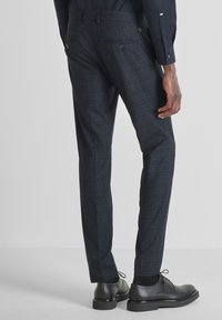 Muškarac u tamnoplavim kariranim hlačama, crnim cipelama, crnim čarapama i tamnoplavoj dugoj majici, stoji protiv svijetle pozadine.