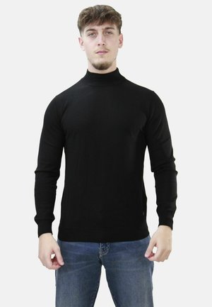 Uomo con capelli corti e ricci che indossa un maglione nero a collo alto a maniche lunghe e jeans blu, in piedi davanti a uno sfondo semplice.