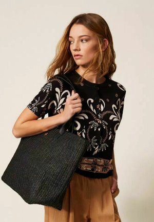 Giovane donna con capelli lunghi che indossa una maglia nera con stampa floreale e pantaloni marrone chiaro, che porta una grande borsa nera dalla texture evidente.