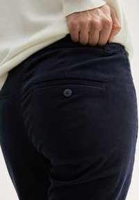 Marineblauwe corduroy broeken hebben een slim fit, achterzakken met zwarte knopen en een gestructureerde stof, gedeeltelijk verborgen onder een crèmekleurige trui.