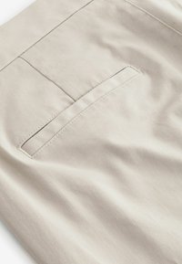 Beige puuvillakangas, joka esittelee sileää pintaa, jossa on ommeltu tasku. Suunnittelussa on hienovaraisia muotoja ja klassinen, räätälöity malli.