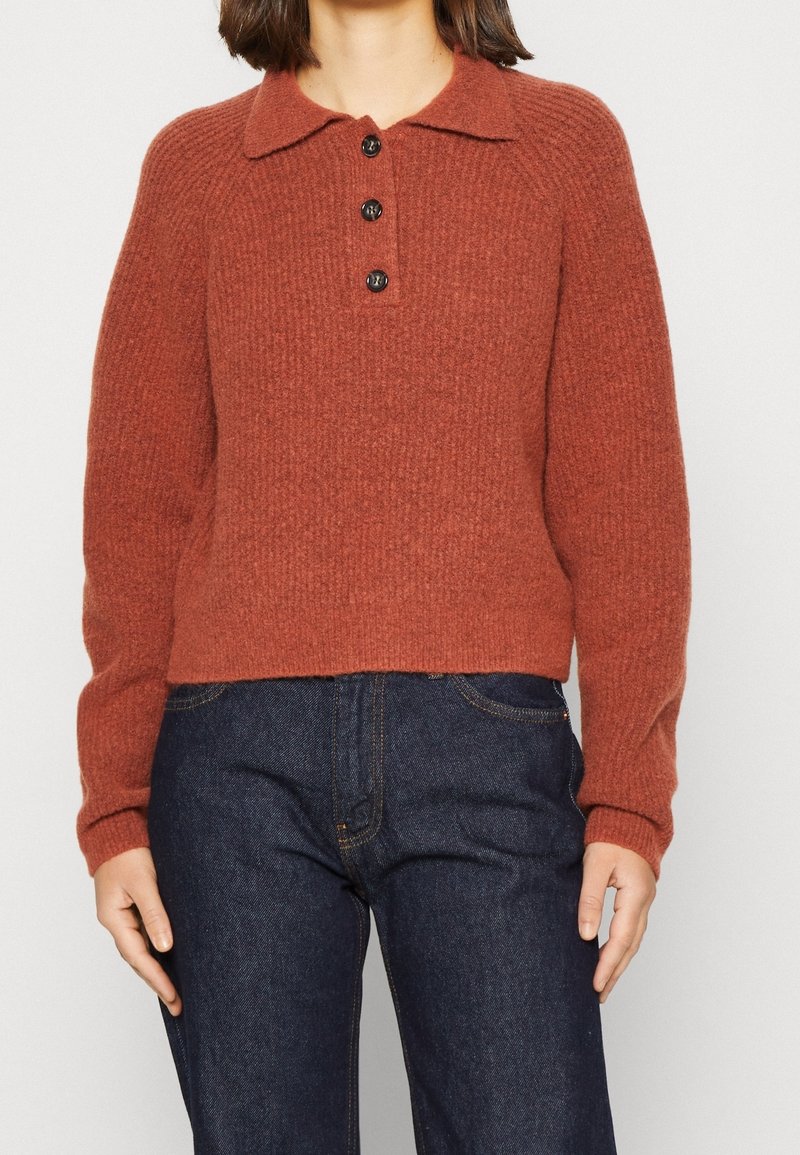 Brede trui in roestkleur met een kraag, voorzien van een knopenstrook en een geribbelde textuur. Gecombineerd met donkere spijkerbroek.
