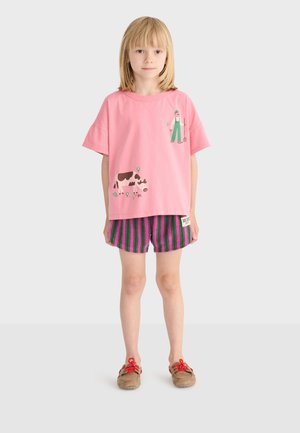 Enfant debout face à l'avant, portant un t-shirt rose avec des illustrations d'agriculteur et de vache, un short rayé violet et vert, et des chaussures marron avec des lacets rouges.