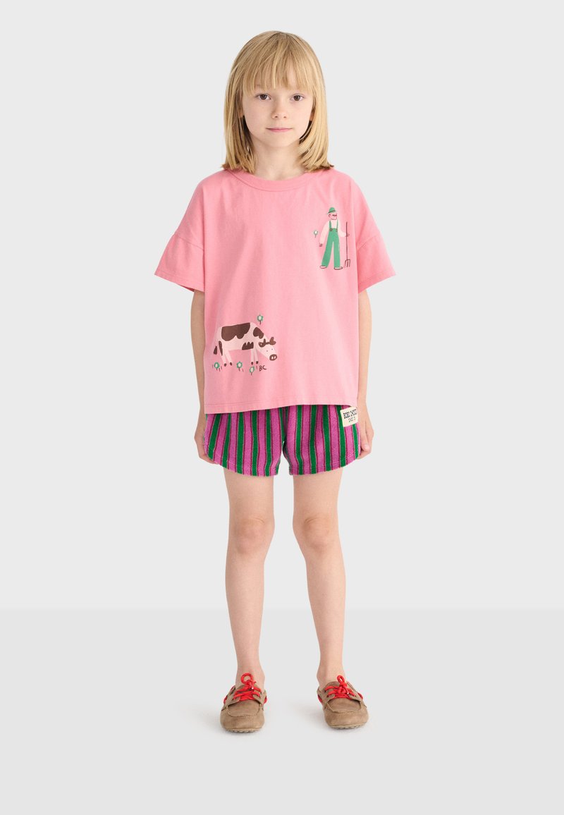 Enfant debout face à l'avant, portant un t-shirt rose avec des illustrations d'agriculteur et de vache, un short rayé violet et vert, et des chaussures marron avec des lacets rouges.