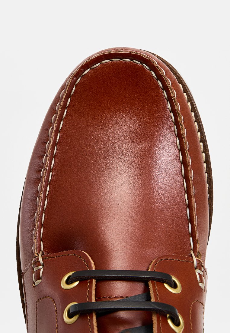 Brązowy skórzany but żeglarski z przodu, z białymi szwami i czarnymi sznurowadłami przeciągniętymi przez złote oczka na jasnym tle.