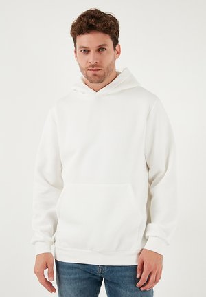 Valkoinen huppari, jossa on etutasku, pitkät hihat ja kiristysnaru hupussa. Sileä pinta ja yksinkertainen muotoilu. Päällä siniset farkut.