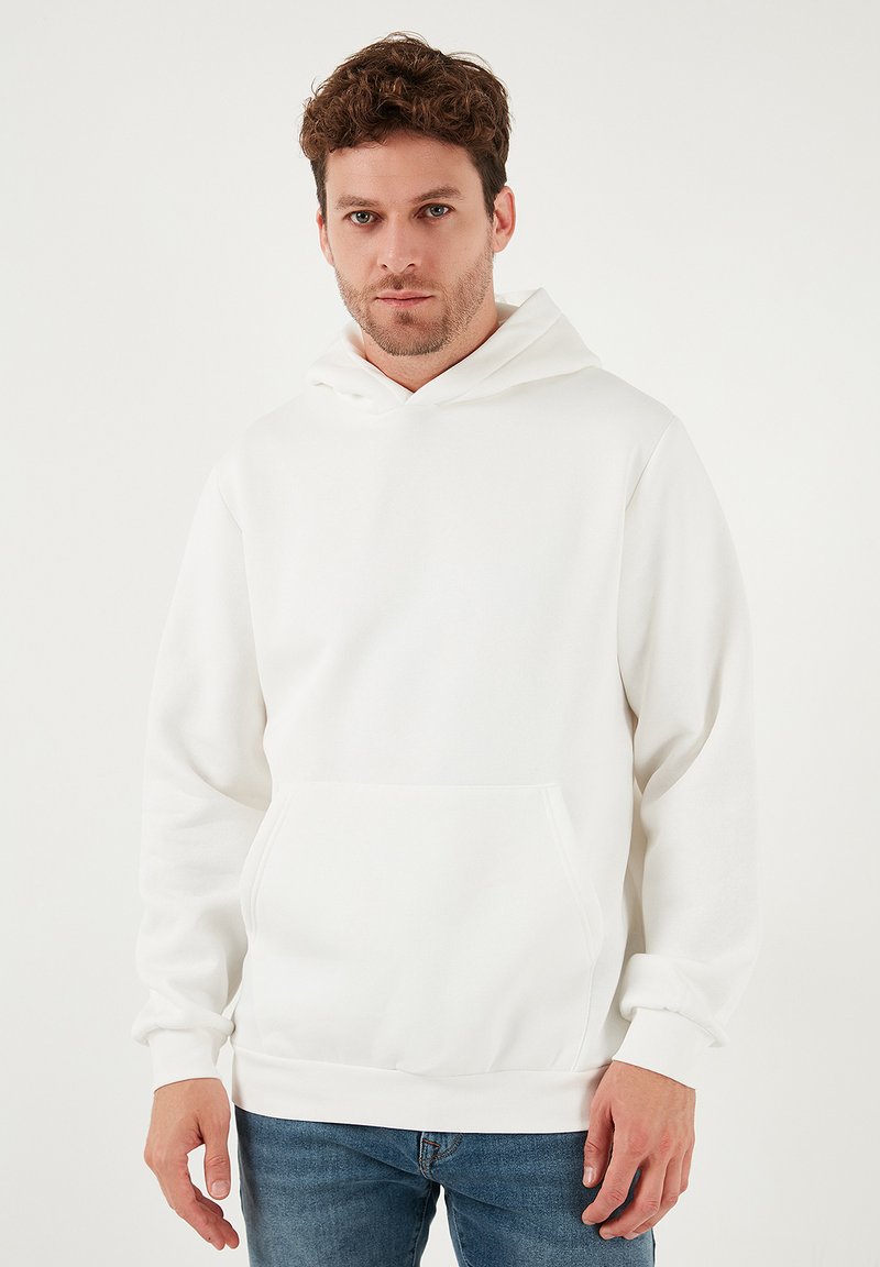 Valkoinen huppari, jossa on etutasku, pitkät hihat ja kiristysnaru hupussa. Sileä pinta ja yksinkertainen muotoilu. Päällä siniset farkut.