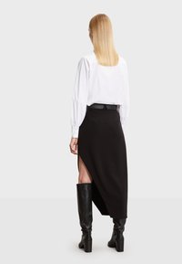 Crna suknja s bočnim prorezom, uparena s bijelom košuljom dugih rukava. Model nosi visoke crne čizme, ističući glatku, teksturiranu tkaninu.