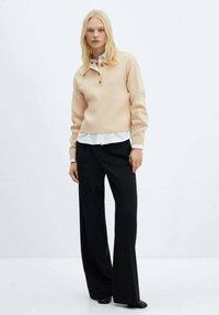 Beiger Strickpullover mit Kragen und drei Knöpfen, über ein weißes Hemd getragen. Kombiniert mit schwarzen, weit geschnittenen Hosen.